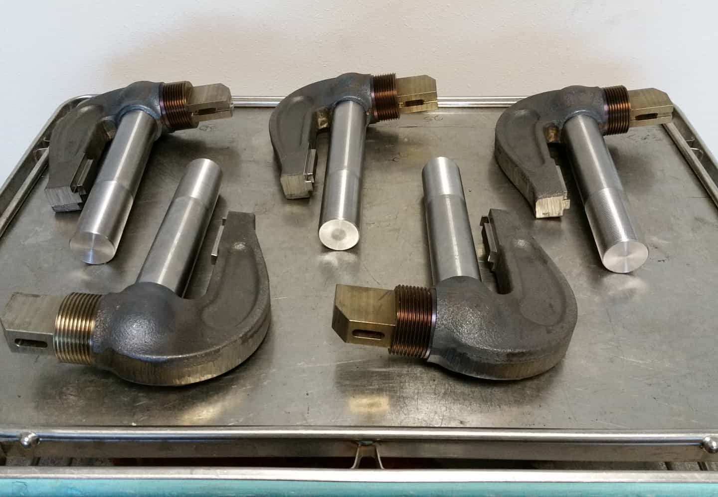 Five freshly machined steel clamp assemblies with threaded shafts lined up on a steel cart